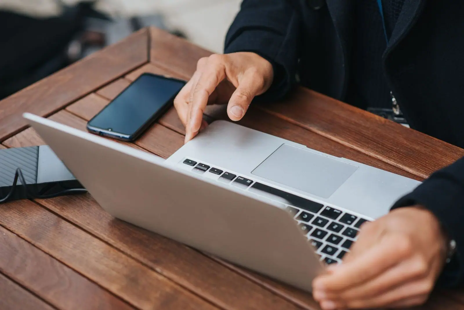 A person at an outdoor table uses a laptop while a smartphone lies beside the keyboard.