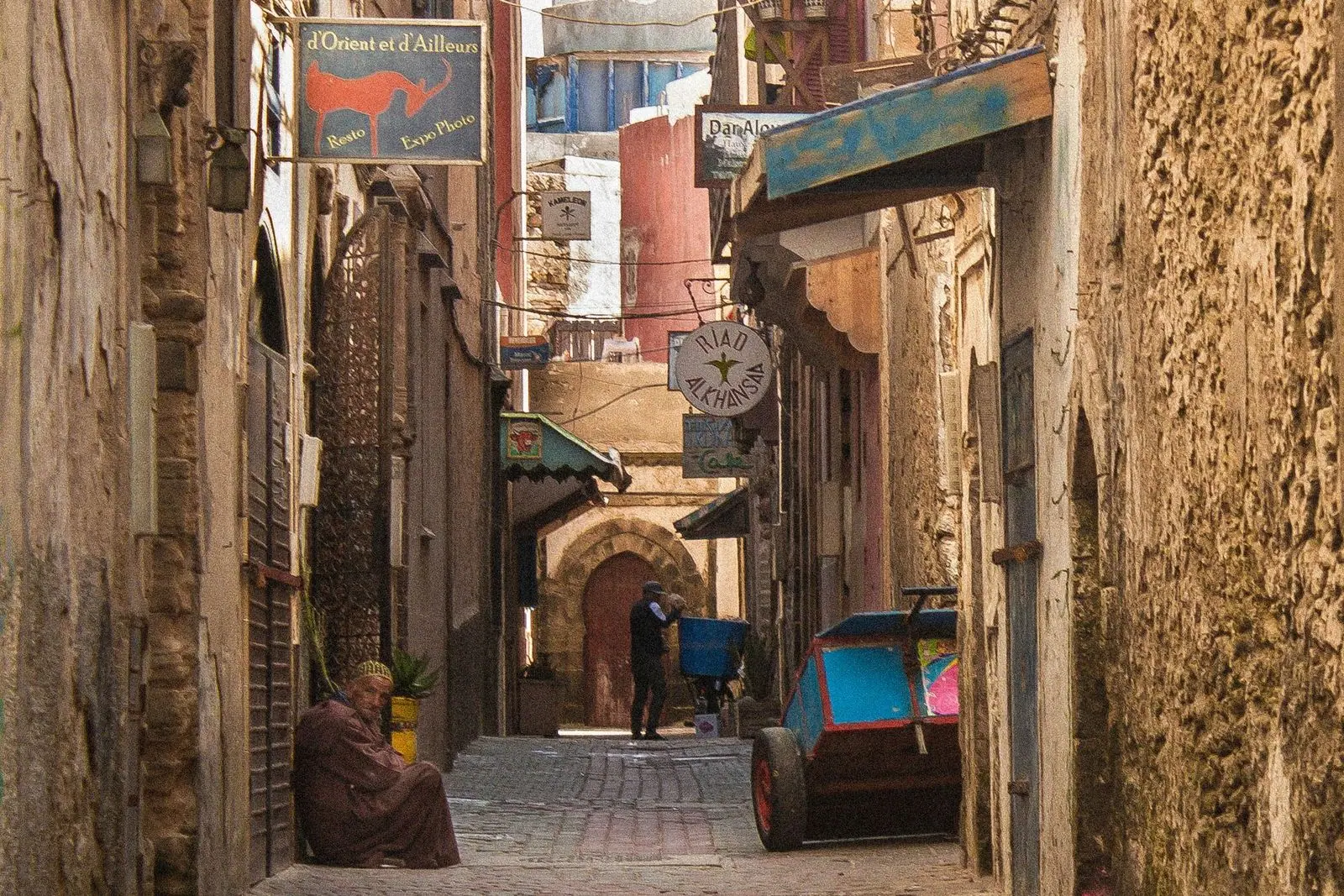 A person sits in a narrow Essaouira alley beside stone walls and painted shop signs.