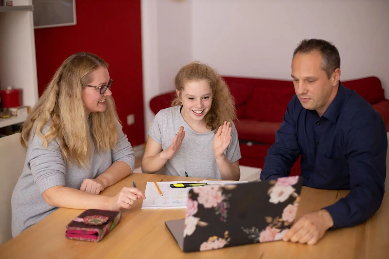 Three people sit at a wooden table, a girl waving toward a laptop while two adults look on.