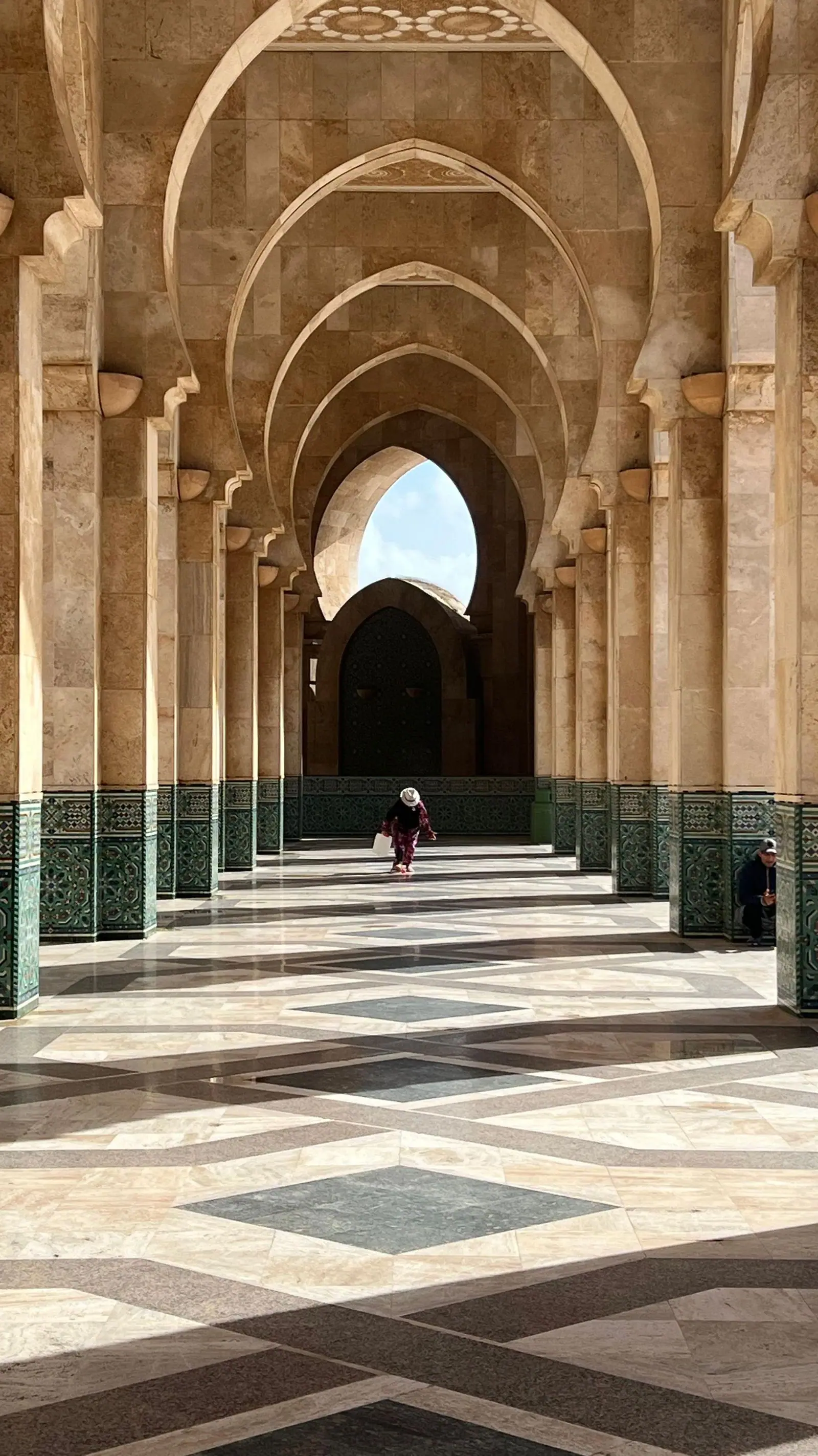 A stone colonnade of repeated arches inside the Hassan II Mosque, with a person walking on the patterned floor.