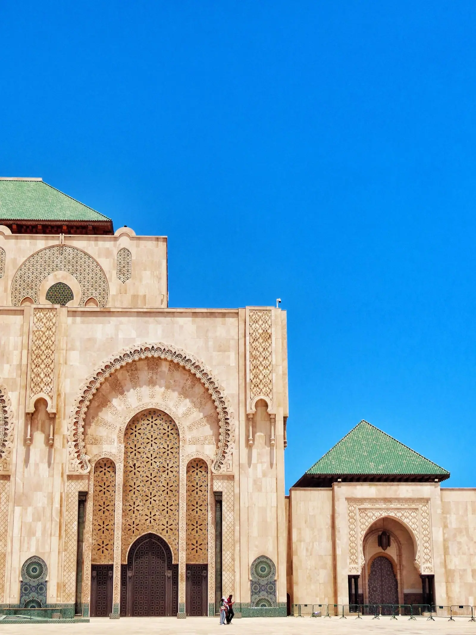 The carved stone façade and green-tiled roof of the Hassan II Mosque in Casablanca, with two people standing in the courtyard.